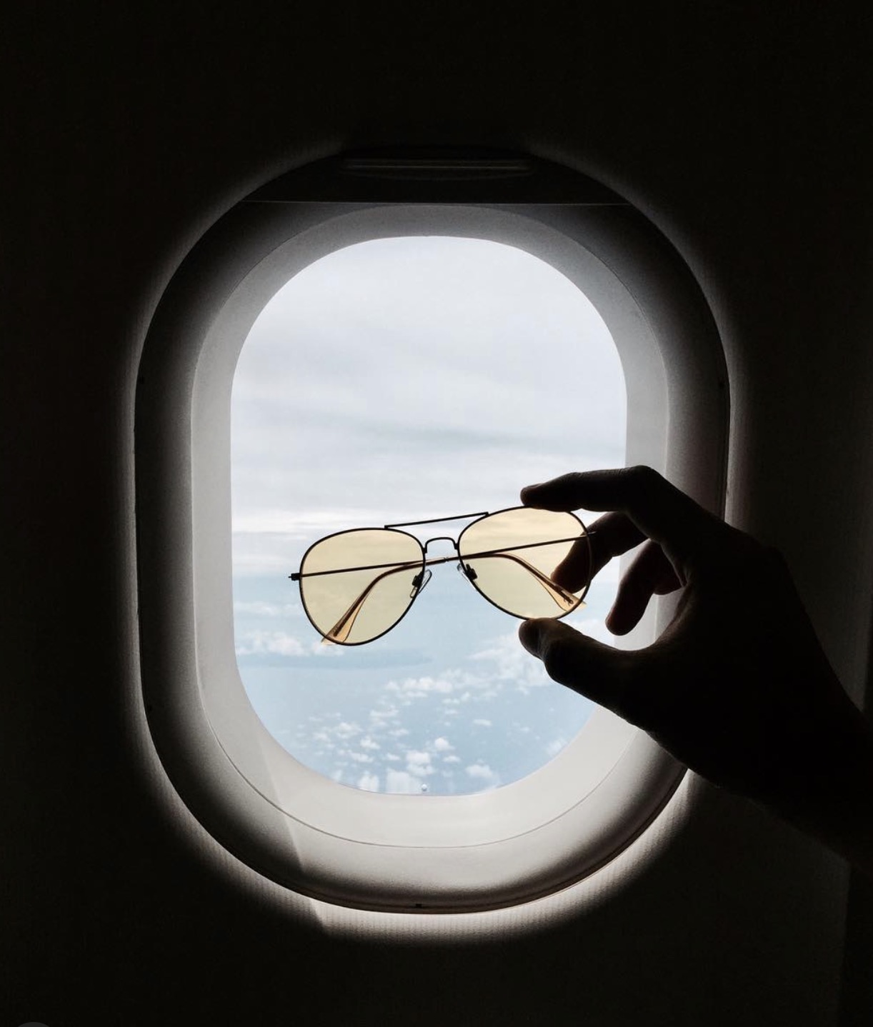 Sunglasses held up to an airplane window overlooking the clouds